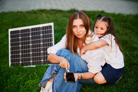 Mother connecting smartphone to photovoltaic solar panel for charging, explains its workings to daughter, sit on grass. Integration of sustainable renewable energy into everyday life.の写真素材