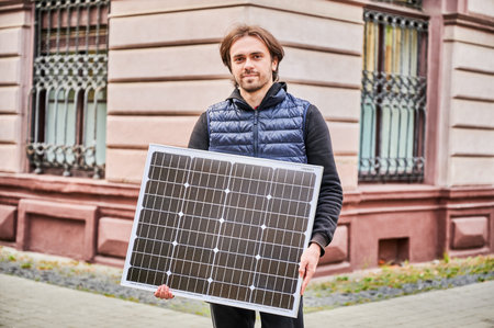 Man holding solar panel, stands in front of old building. Guy in a black hoodie and navy blue quilted vest. Aged architecture contrasts with modernity of renewable energy.の写真素材