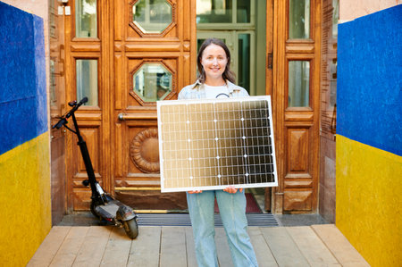 Smiling woman holding solar panel, emphasizing integration of sustainable renewable energy. Electric scooter parked nearby, with colorful blue and yellow colors of Ukrainian flag walls flanking scene.の写真素材