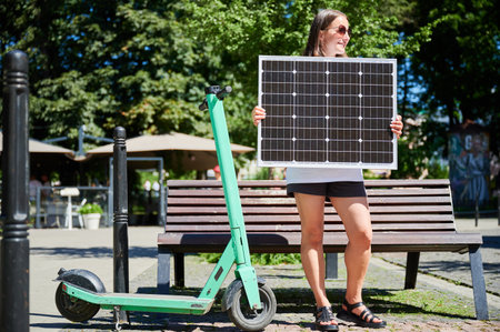 Happy woman charging her electric scooter from solar panel. Young female holding solar battery and catching sunlight. Concept of renewable energy resources.の写真素材