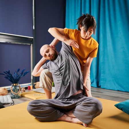 Masseuse making thai yoga massage. Woman therapist doing traditional massage treatment, stretching male patients body. Bald client sitting on mat, enjoying hand massage in calm atmosphere.の写真素材