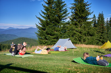 Group of people doing yoga pose outdoor in camping in the mountains. Adults and children on yoga mats, each doing a yoga pose under a clear blue sky in the morning. Young boy is instructor.の写真素材