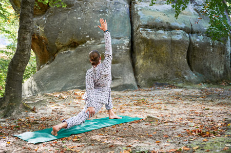 Woman practicing yoga outdoors in forest. Barefoot female on yoga mat surrounded by trees and large rocks, which suggests peaceful, natural environment ideal for meditation or yoga practice.の写真素材