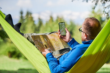 Man traveler in glasses studies map, checks his phone, combining traditional and modern navigation methods planning outdoor adventure or hiking route, relaxes in green hammock in forested area.の写真素材