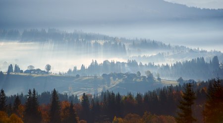 Morning sunlight streams through mist, illuminating serene mountain village nestled among forested hills. Light creates ethereal rays that gently touch autumn foliage and houses scattered on slopes.の写真素材