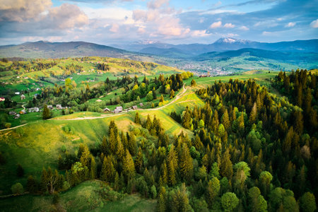 Aerial view of picturesque rural landscape in Ukraine, with lush green fields, dense forests, and scattered houses under sky filled with dramatic clouds. Snow-capped mountains in distant horizon.の写真素材