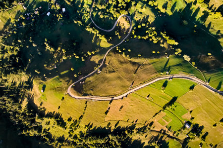 Top-down aerial view of picturesque rural landscape bathed in golden sunlight. Winding roads meander through green fields and small patches of trees. Scattered farmhouses and haystacks dot terrain.の写真素材