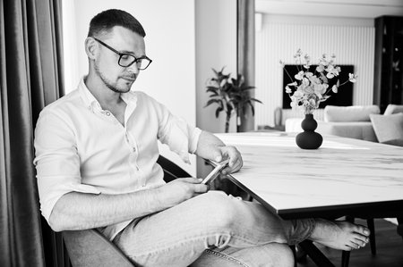 Portrait of man engaging with smartphone sitting at white marble countertop on kitchen, wearing glasses. Handsome man relaxing in his minimalist and modern aesthetic apartment.の写真素材