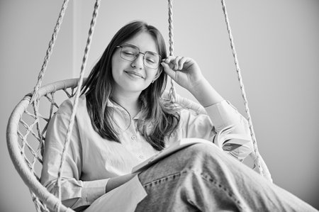 Portrait of girl relaxing with book on indoor swing with closed eyes. Pretty female in glasses excited about story she reading. Cozy afternoon with book in hanging chair in home. Black and white imageの写真素材