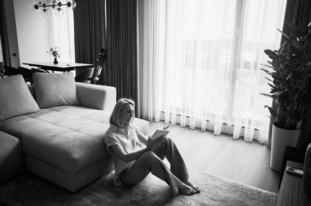 Young woman sitting next to couch and reading. Beautiful female relaxing on wooden floor. Woman sitting with book opposite window in natural light. Black and white image.の写真素材