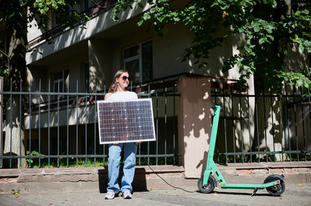 Woman using portable solar panel to charge scooter, highlighting integration of sustainable renewable energy. Green electric scooter connected to solar panel, emphasizing eco-friendly transportation.の写真素材