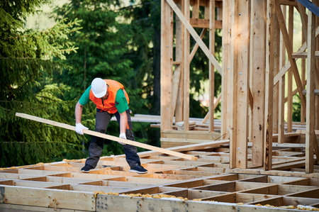 Carpenter constructing wooden-framed house near the woods at sunset. Man holding truss and dressed...