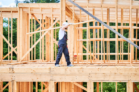 Carpenter constructing wooden-framed house near. Man holds large beam on his shoulder and is dressed in work clothes and helmet. Modern and environmentally-friendly construction concept.の写真素材