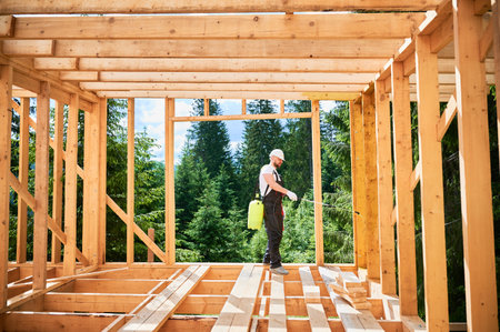 Laborer constructing wooden frame house near forest. Man treating woods, applying fire retardant using sprayer, while dressed in protective suit, helmet. Concept of modern eco-friendly construction.の写真素材