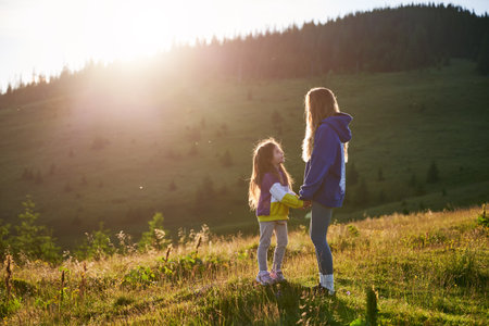 Young woman tourist spending time together with little daughter in sunlit meadow, against stunning mountain backdrop at sunset. Joyful moment captures essence of carefree fun and natural beauty.の写真素材