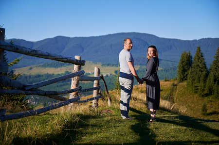 Couple walks hand-in-hand along rustic fence. Man and woman enjoying stunning mountain landscape. Clear blue sky and lush greenery create peaceful and romantic atmosphere.の写真素材