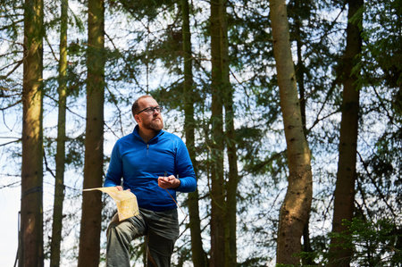 Man traveler in glasses stands in dense forest, holding map in one hand and compass in other. Tourist planning outdoor adventure or hiking route, amidst tall trees and natural surroundings.の写真素材