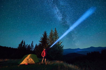 Man hiker stands under dazzling starry sky, shining flashlight into heavens. Milky Way stretches above, glowing tent nearby, creating magical and serene scene in peaceful mountain wilderness.の写真素材
