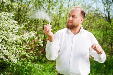 Man allergic using medical nasal drops, suffering from seasonal allergy at spring in blossoming garden. Handsome man examining nasal spray near blooming tree outdoors. Spring allergy concept.の写真素材