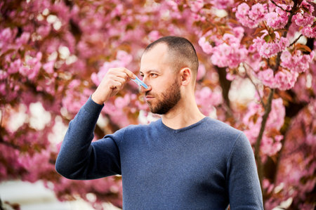 Man allergic suffering from seasonal allergy at spring. Bearded man with clothespin clipped to his nose - symbolic gesture of his inability to breathe due to nasal congestion near blooming tree.の写真素材