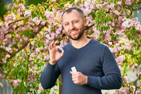 Man allergic suffering from seasonal allergy in blossoming garden at spring. Handsome smiling man holding allergy drug, making OK hand sign in front of blooming tree. Spring allergy concept.の写真素材