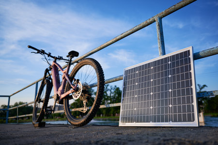 Focus on solar panel against backdrop of blue sky and distant trees. Pink electric mountain bike parked by lakeside railing, connected to solar panel for charging.の写真素材