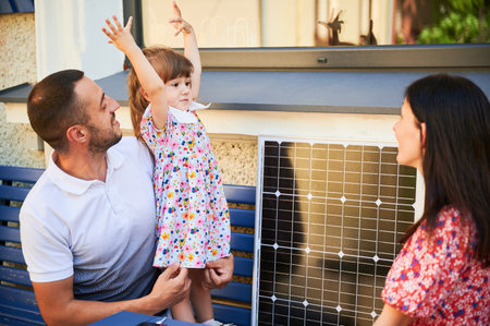 Happy family spending time together with solar panel. Father, holding young girl, sits on bench with mother. Scene highlights importance of educating next generation about renewable energy.の写真素材