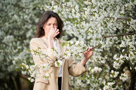 Woman allergic suffering from seasonal allergy at spring in blossoming garden at springtime. Young woman sneezing, having runny nose in front of blooming tree. Spring allergy conceptの写真素材