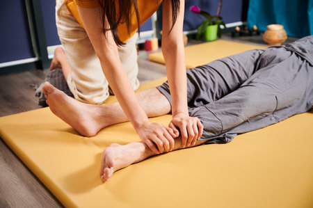 Masseuse making thai yoga massage. Cropped view of woman therapist doing traditional massage treatment for male patient. Client lying on mat, meditating, enjoying hand massage in calm atmosphere.の写真素材