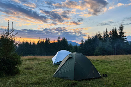 Few tourist tents in grassy field in evening, surrounded by trees and hills under stunning sunset sky. Colorful clouds enhance beauty of scene, with hues of orange and purple illuminating horizon.の写真素材