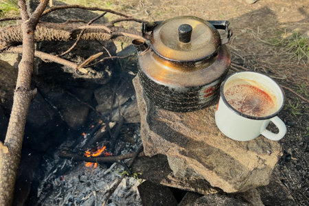 White enamel mug filled with hot beverage sits beside rustic black kettle on stone fire pit. Nearby, some sticks arranged for fire, all set against a backdrop of a green, forested camping area.の写真素材