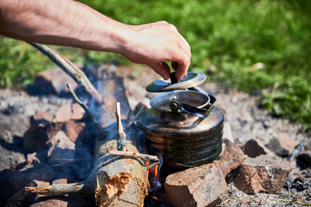 Close-up shot of hand lifting lid off blackened kettle heating over campfire. Kettle on ring of stones with flames and smoke rising around it. Grassy meadow, emphasizing outdoor camping experience.の写真素材