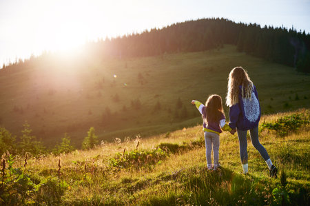 Young woman tourist spending time together with little daughter in sunlit meadow, against stunning mountain backdrop at sunset. Joyful moment captures essence of carefree fun and natural beauty.の写真素材