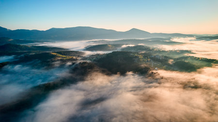 Aerial view reveals misty morning landscape with rolling hills and villages. Golden sunlight breaks through fog, illuminating patches of green and creating dreamy, ethereal scene under clear blue sky.の写真素材