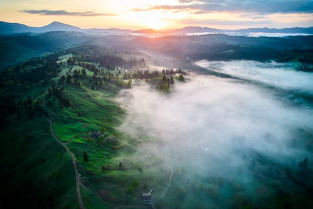 Aerial view of sunrise over mist-covered landscape, with golden light illuminating rolling hills and valleys. Thick fog creates mystical atmosphere, partially obscuring lush greenery and trees below.の写真素材