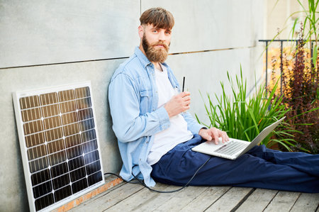 Portrait of barded man works on laptop on lap with glass of iced coffee. Solar panel nearby, emphasizing sustainable, modern eco-friendly workspace that combines technology and sustainability.の写真素材