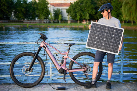 Man cyclist using solar panel for charging electric mountain bike outdoors. Guy in helmet and sunglasses stands by lake, enjoying serene waterside scenery on sunny evening.の写真素材