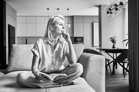 Young woman thoughtfully looking away. Beautiful female relaxing on couch and reading. Woman in casual clothes sitting on cream sofa and thinking. Black and white image.の写真素材