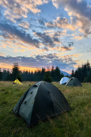 Few tourist tents in grassy field in evening, surrounded by trees and hills under stunning sunset sky. Colorful clouds enhance beauty of scene, with hues of orange and purple illuminating horizon.の写真素材