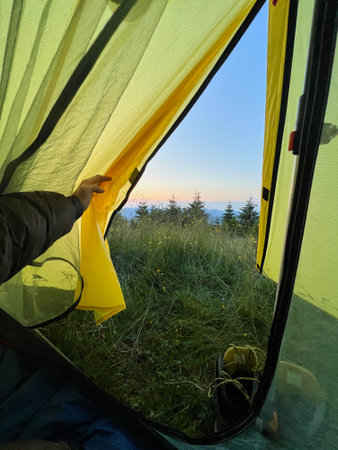 Camper peeks out from yellow tent at dawn, revealing serene view of grassy terrain and distant trees. Dewdrops glisten on tent fabric, with hiking boots placed at tents entrance.の写真素材