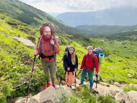 Three hikers, dressed in colorful outdoor gear, pause on rocky, green mountain trail. Scenic backdrop features lush hills and partly cloudy sky.の写真素材