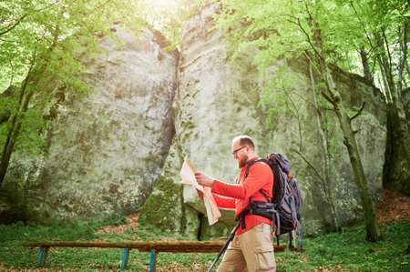Happy bearded tourist man studies map while stands in front of large rock in dense forest. Smiling traveler with grey backpack and glasses, planning outdoor adventure, hiking or climbing route.の写真素材