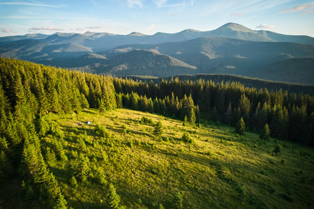 Aerial view tent in sunlit grassy clearing, surrounded by dense forest. Expansive scene reveals rolling green hills and towering mountains under clear blue sky, creating idyllic, serene camping spot.の写真素材