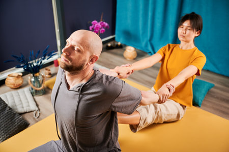 Masseuse making thai yoga massage. Woman therapist doing traditional massage treatment, stretching male patients body. Bald client sitting on mat, enjoying hand massage in calm atmosphere.の写真素材