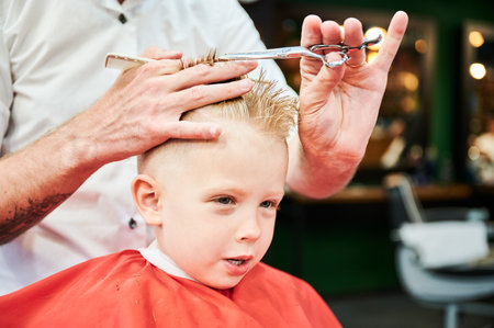 Man barber cutting little boys hair using comb and scissors. Child getting haircut from adult male, likely barber. Professional hairdresser and cute client at modern barbershop.の写真素材