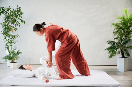 Masseuse making thai yoga massage. Woman therapist doing traditional massage treatment, stretching female patients body. Client lying on mat, enjoying hand massage in calm atmosphere.の写真素材