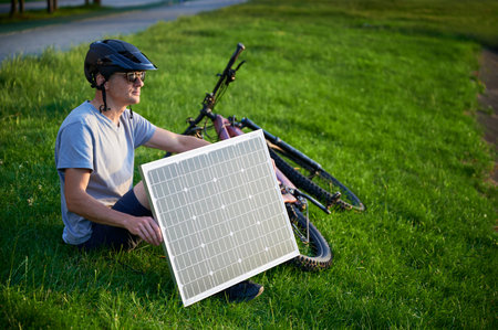 Man cyclist using solar panel for charging electric mountain bike outdoors. Guy wearing helmet and sunglasses sits on grass in park, enjoying sunny evening.の写真素材