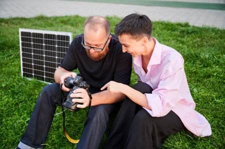 Couple sits on grass, closely examining photos on camera, with solar panel nearby. Integration of...