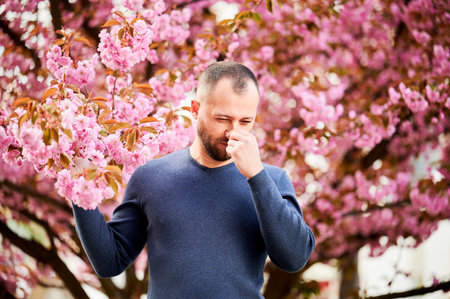 Man allergic suffering from seasonal allergy at spring in blossoming garden at springtime. Bearded young man sneezing and having runny nose in front of blooming tree. Spring allergy concept.の写真素材