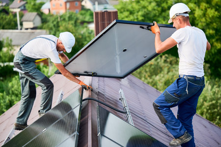 Workers building solar panel system on roof of house. Two men installers in helmets carrying photovoltaic solar module outdoors. Alternative, green and renewable energy generation concept.の写真素材